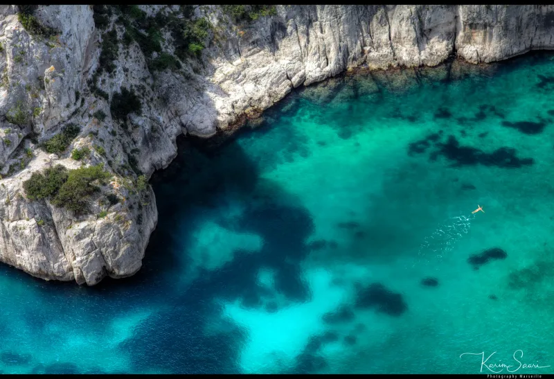Vue aérienne plongeante sur une calanque turquoise, falaises calcaires blanches, silhouette d'un nageur dans l'eau