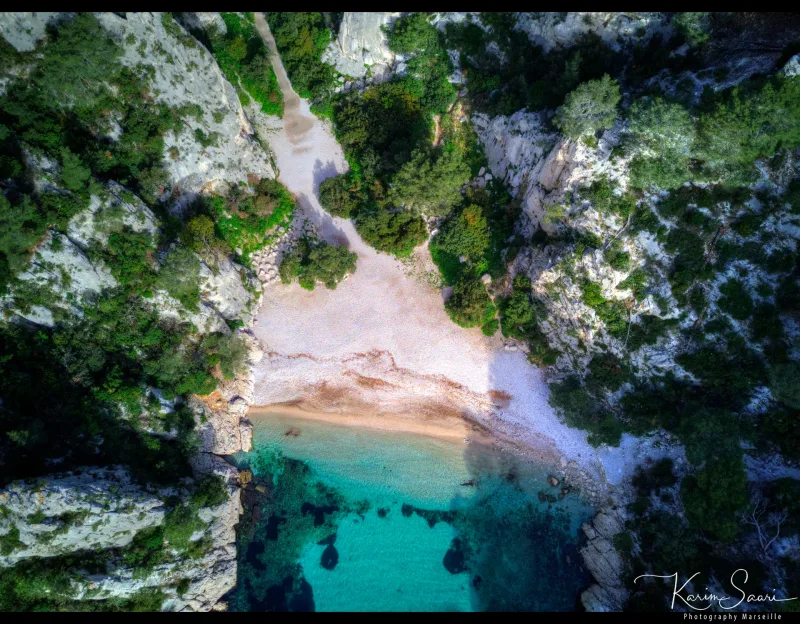 Vue aérienne d'une calanque secrète, plage de galets, eau émeraude encadrée de falaises calcaires et de végétation