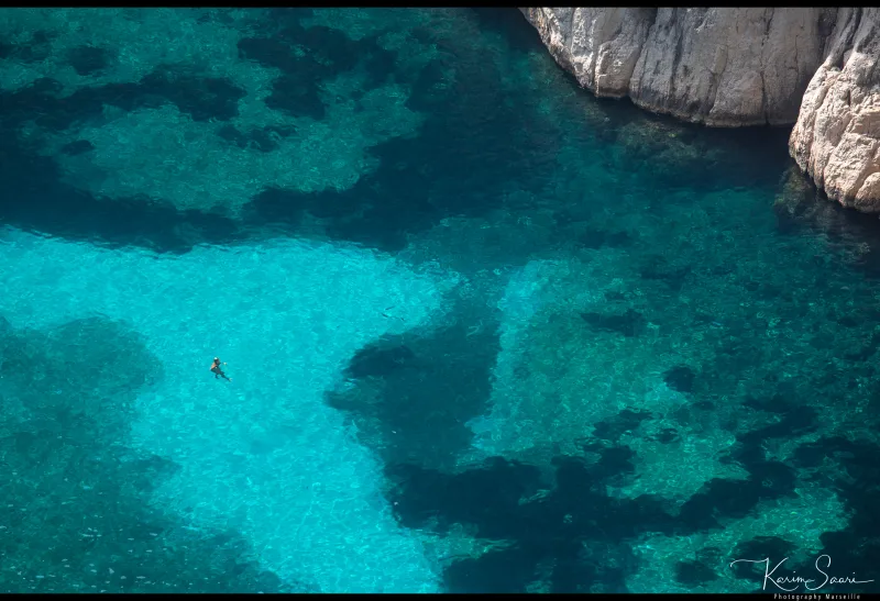 Vue aérienne plongeante sur un nageur solitaire dans les eaux turquoise d'une calanque entre rochers calcaires