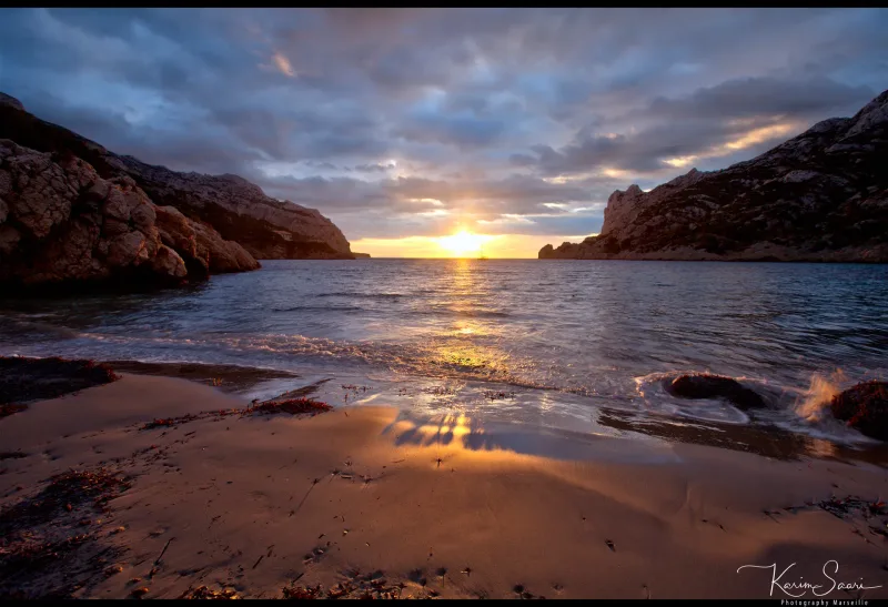 Coucher de soleil sur une plage de calanque entre deux falaises, ciel dramatique, reflets dorés sur le sable mouillé