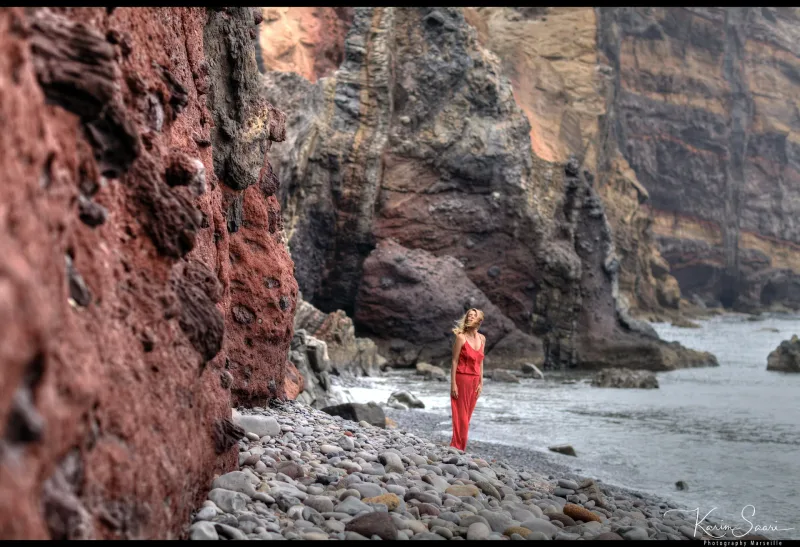Femme en robe rouge sur une plage de galets face aux falaises volcaniques rouges — paysage côtier Marseille