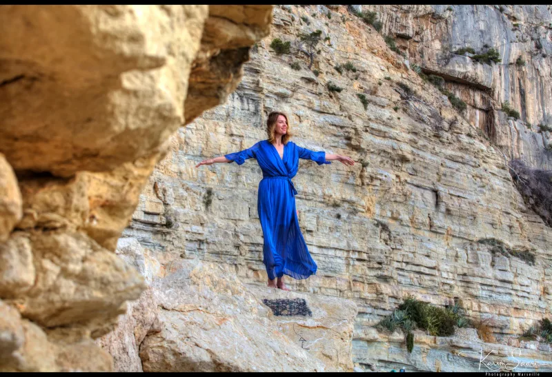 Femme en robe bleue les bras écartés sur un rocher calcaire en strates dans les Calanques de Marseille
