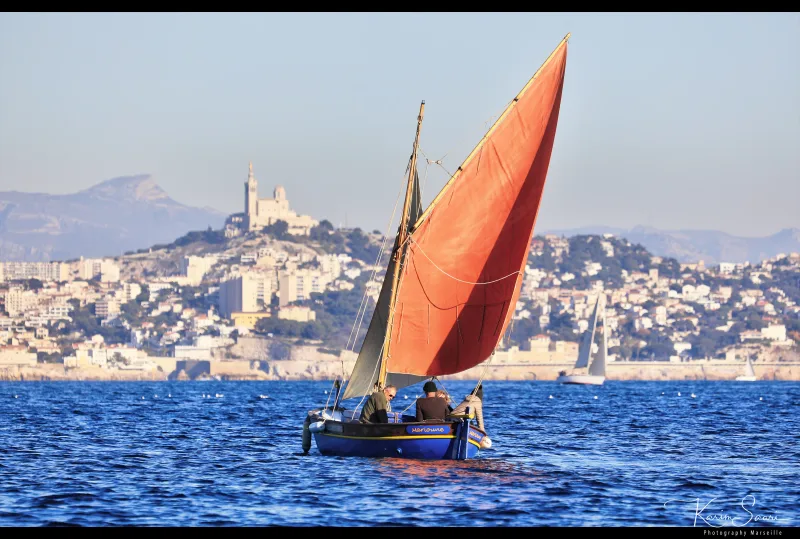 Pointu marseillais à voile rouge naviguant devant Marseille avec Notre-Dame de la Garde en arrière-plan