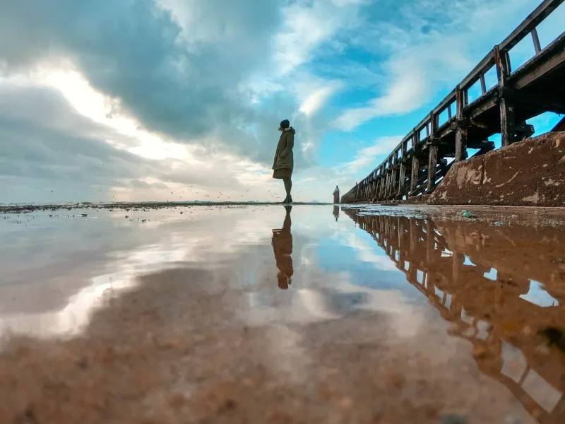 Silhouette d'une personne se reflétant dans une flaque sur la plage à côté d'une jetée en bois, ciel dramatique