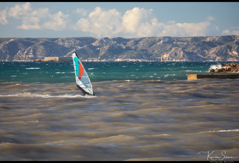 Véliplanchiste sur la mer agitée avec les Calanques de Marseille et le fort en arrière-plan