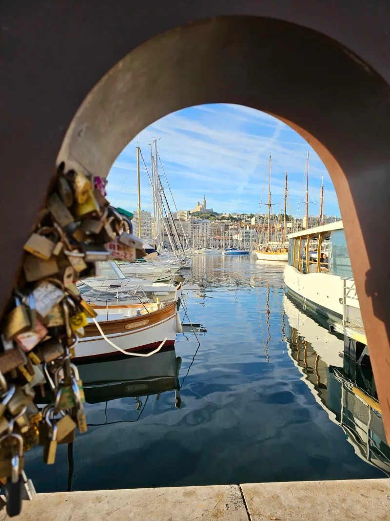 Vieux-Port de Marseille vu à travers une arche ornée de cadenas d'amour, voiliers et Notre-Dame de la Garde