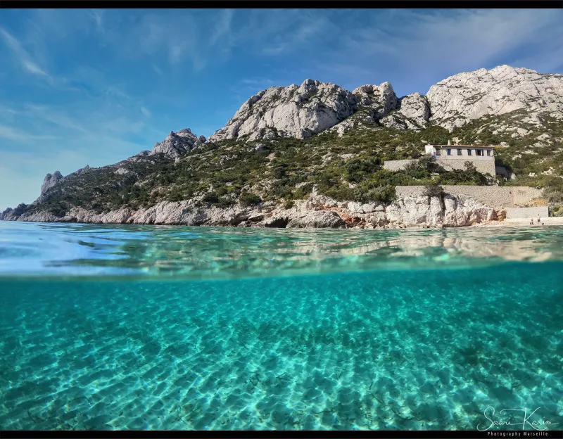 Vue mi-eau mi-ciel d'une calanque turquoise, fonds clairs et galets sous l'eau, falaises calcaires et pin — © Karim Saari, photographe sous-marin Marseille